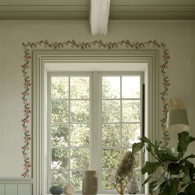 Decorative carnation floral stencil border above a window with a lamp and plant on a light-colored wall.