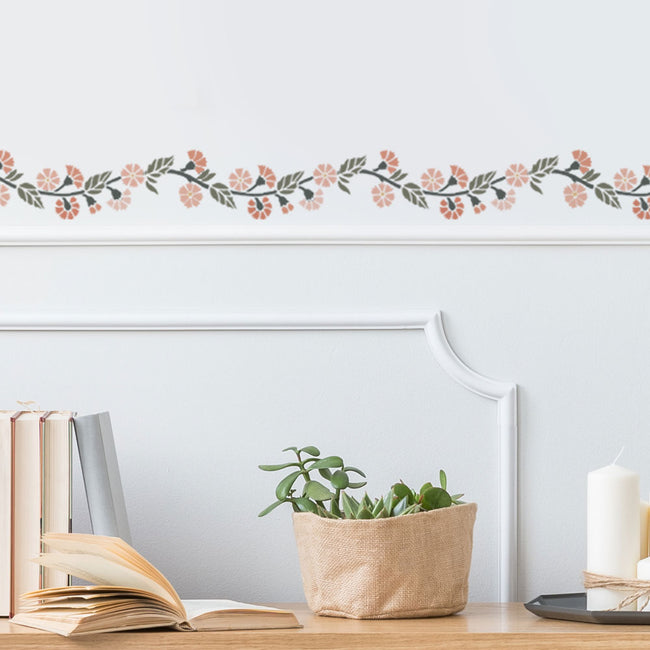 Decorative floral border stencil on a wall with books, plant, and candle on a table.