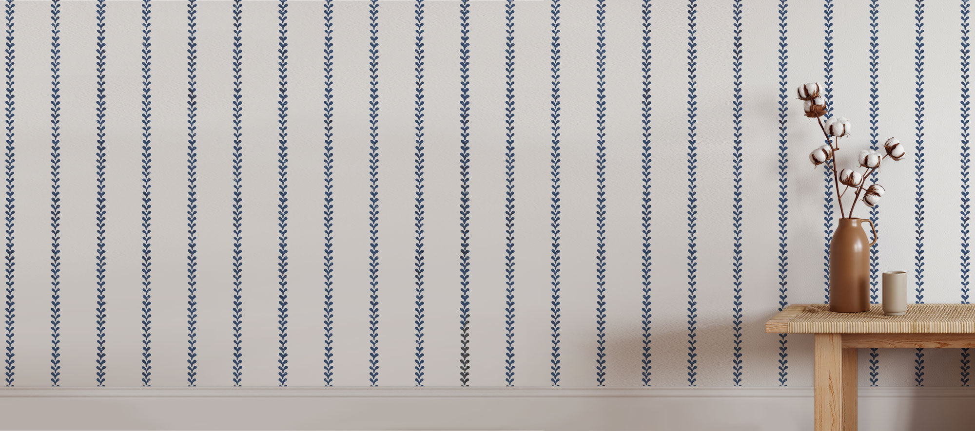 Vertical navy blue and white patterned stencil shown on a wall in a styled room setting, with a small wooden side table and vase holding dried branches on the left.