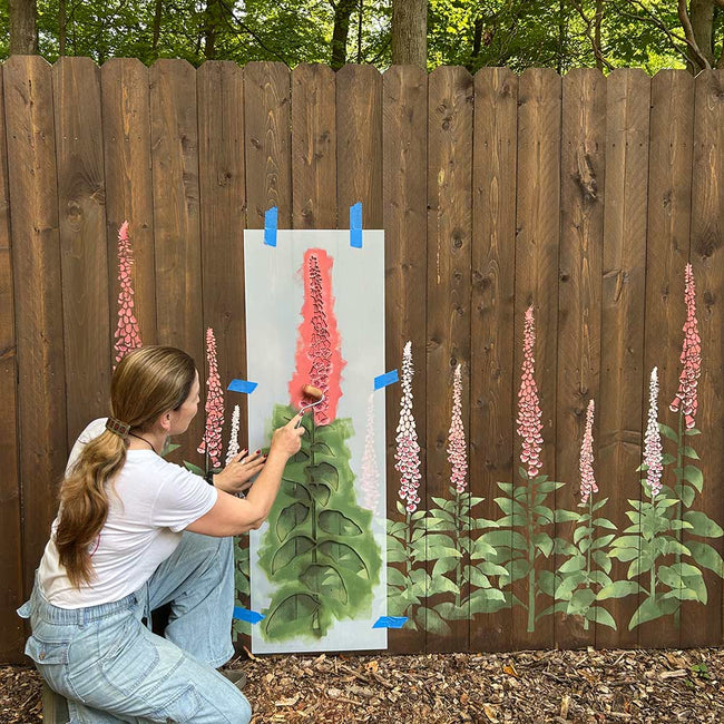 Woman painting wooden fence mural with foxgloves