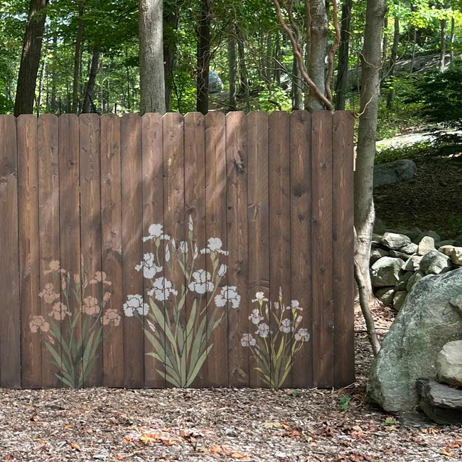 A brown wooden privacy fence with a hand-painted floral design featuring white irises and pink flowers, set in a wooded outdoor environment.