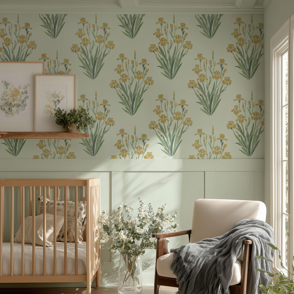 A styled nursery room featuring a stenciled repeating pattern of yellow iris flowers and green leaves on a pale sage green background. The room includes a light wood crib, a cream armchair with a gray throw blanket, a wooden floating shelf with framed botanical prints, and a glass vase of white flowers.