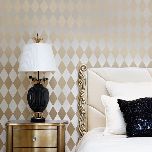 A wall stencil pattern with diamond-shaped designs is shown applied on a wall above a bed with a lamp and a decorative pillow.