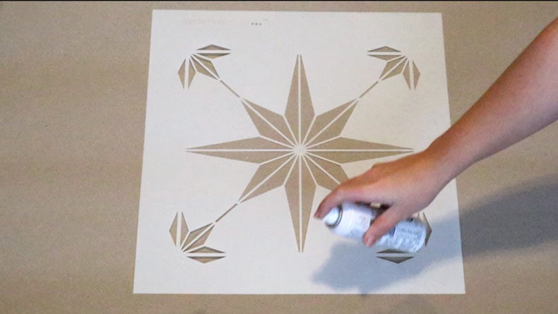 woman's hand spraying spray adhesive on tile stencil over brown paper