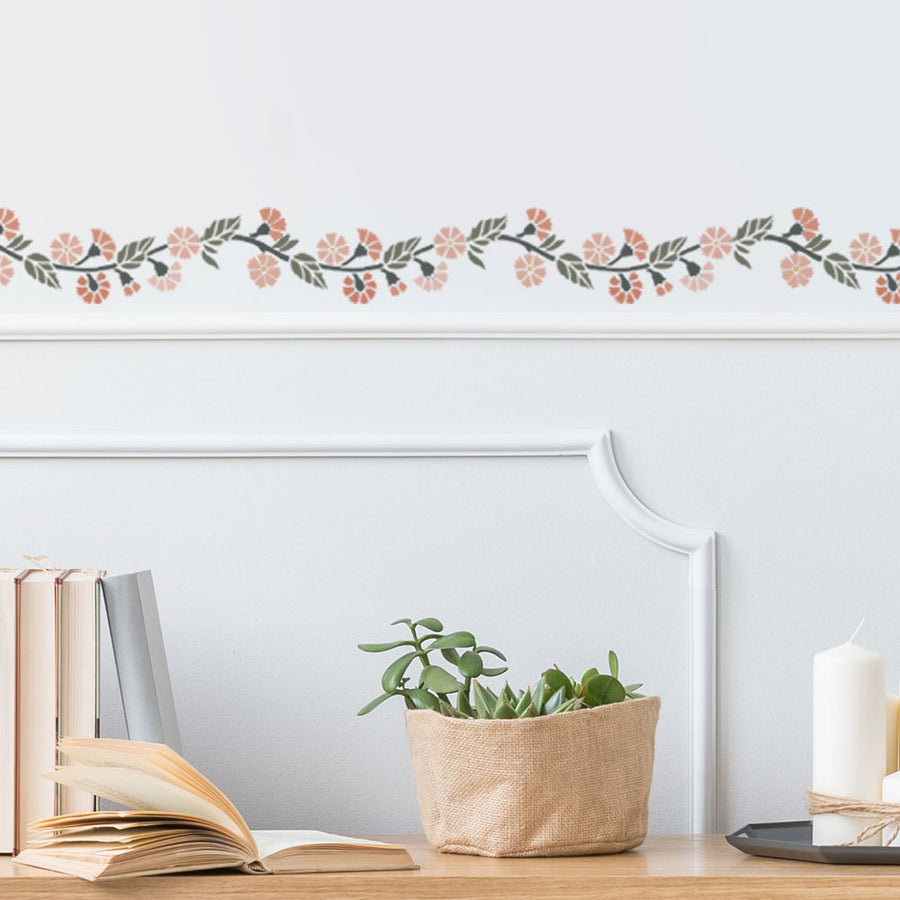 Decorative floral border stencil on a wall with books, plant, and candle on a table.