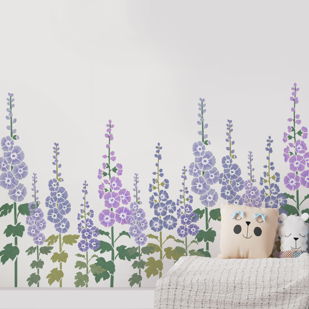 Lavender and purple floral wall stencils with green leaves arranged along a white wall above a bed with a cream bear pillow and a white textured blanket.