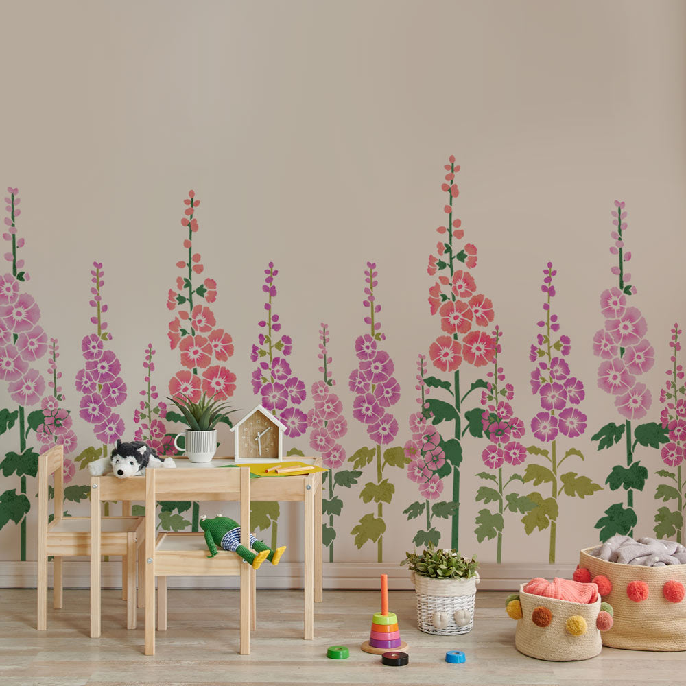A children's playroom corner featuring a stenciled floral wall mural with pink, purple, and red hollyhocks on a beige wall. In front of the wall is a light wood table and chairs with toys, a small potted plant, and storage baskets on the floor.