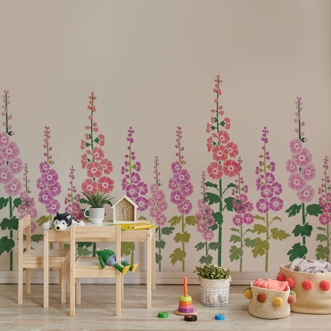 A children's playroom corner featuring a stenciled floral wall mural with pink, purple, and red hollyhocks on a beige wall. In front of the wall is a light wood table and chairs with toys, a small potted plant, and storage baskets on the floor.