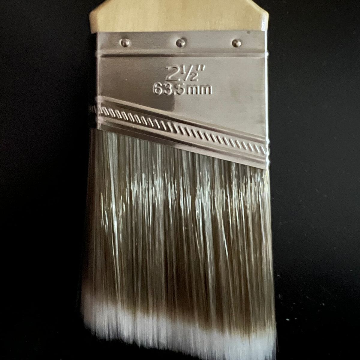 A close-up of a paint brush with a light wood handle, a silver metal ferrule engraved with '2 1/2" 63.5mm', and white bristles on a black background.