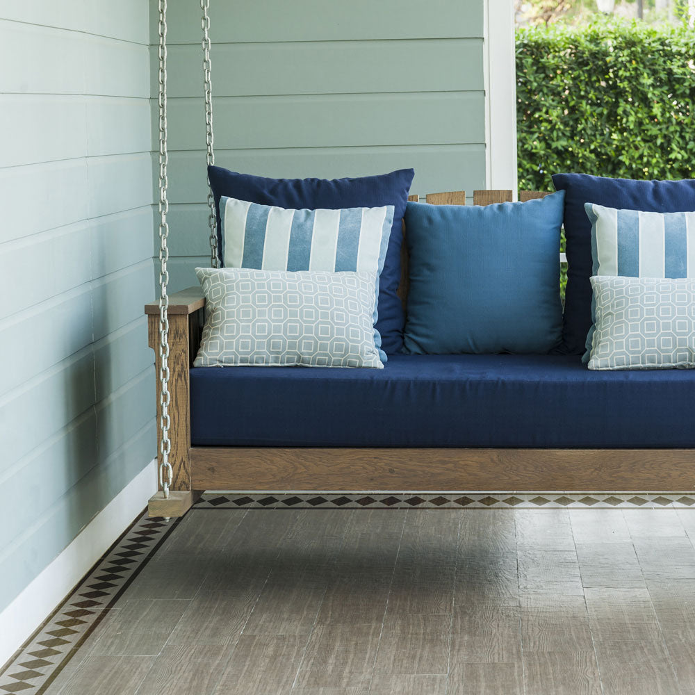 A wooden porch swing daybed with navy blue cushions and decorative throw pillows, set on a stenciled porch with a light green siding wall.