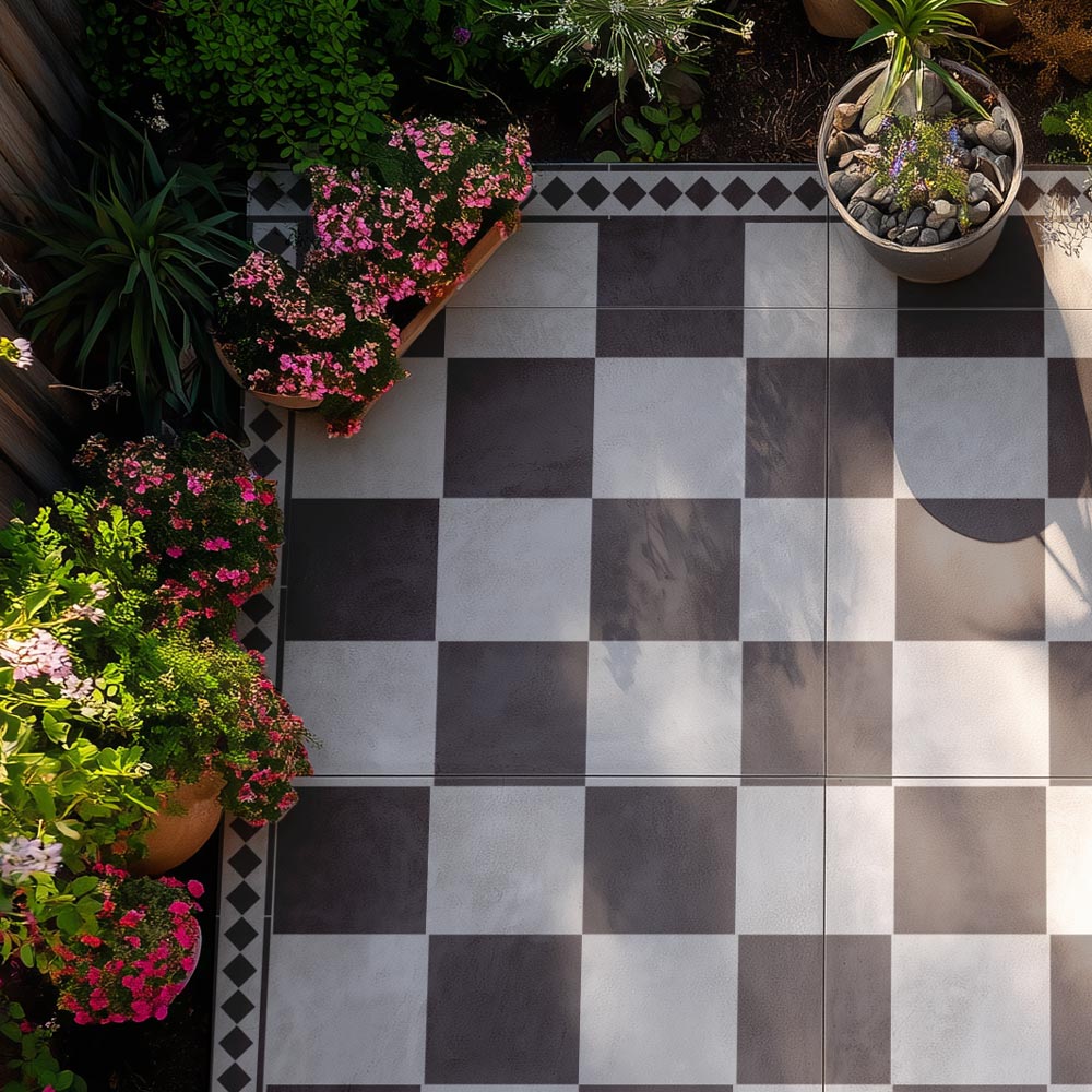 A top-down view of a checkered patio floor with alternating dark charcoal and light gray square stenciled tile, bordered by a black and white diamond-patterned strip. The patio is surrounded by green plants and potted pink flowers.