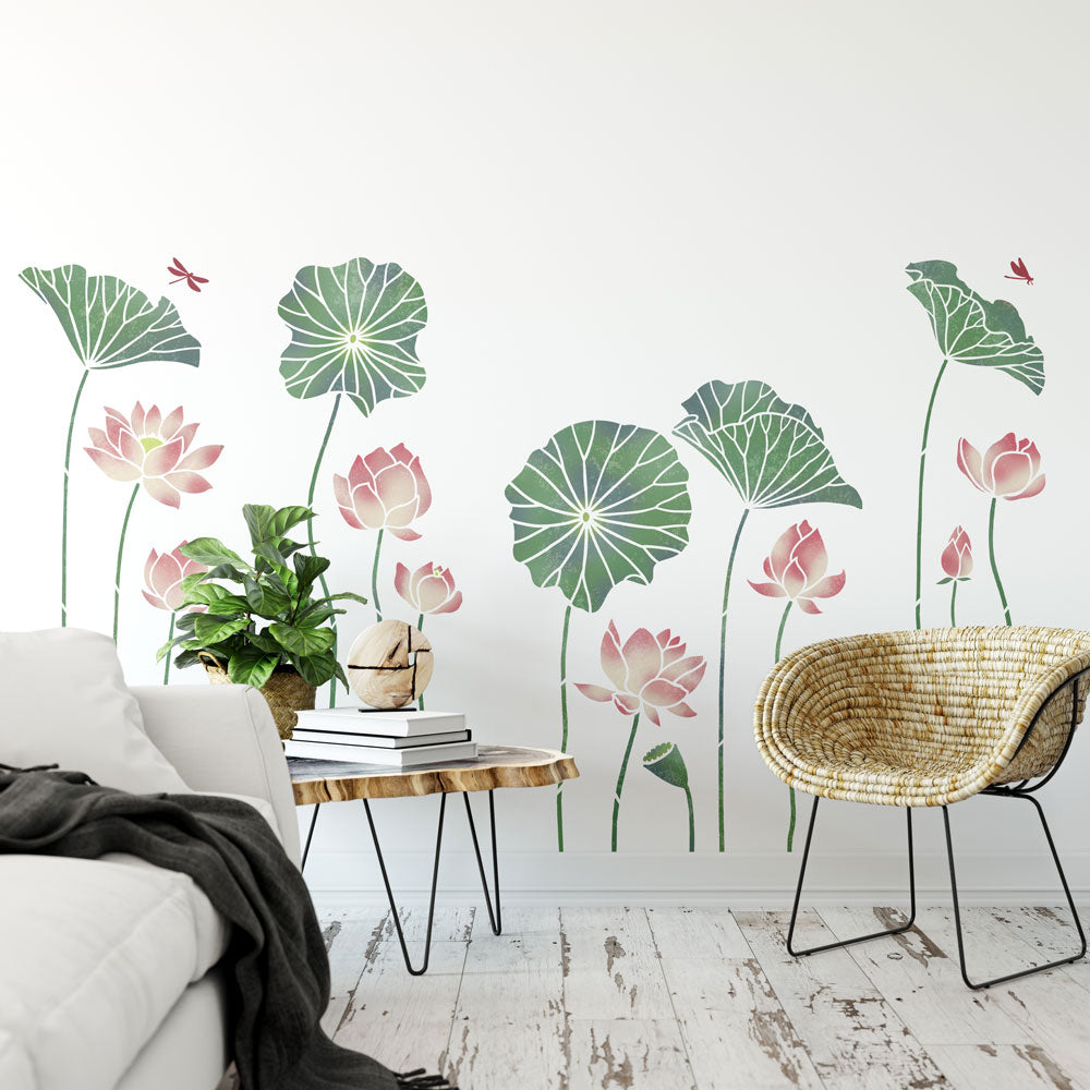 Large-scale wall mural of lotus leaves and pink lotus flowers in a modern living room setting, shown behind a white sofa, a small wooden side table with books and a potted plant, and a woven accent chair.