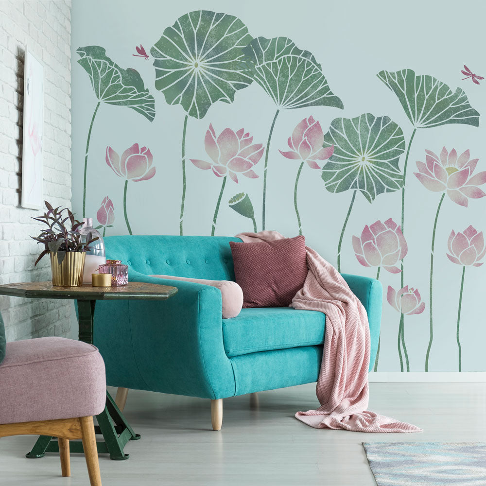 A styled interior scene showing a large wall mural of green lotus leaves and pink lotus flowers on a pale blue wall, with a turquoise armchair, pink throw blanket, side table, and part of a pink chair in the room.
