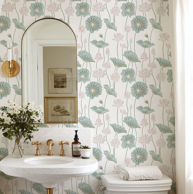 A bathroom interior featuring a wall stencil with a repeating lotus leaf pattern in muted teal and blush pink on a warm off-white background. A white pedestal sink with gold faucets, a wall mirror, framed art, and a toilet are visible.