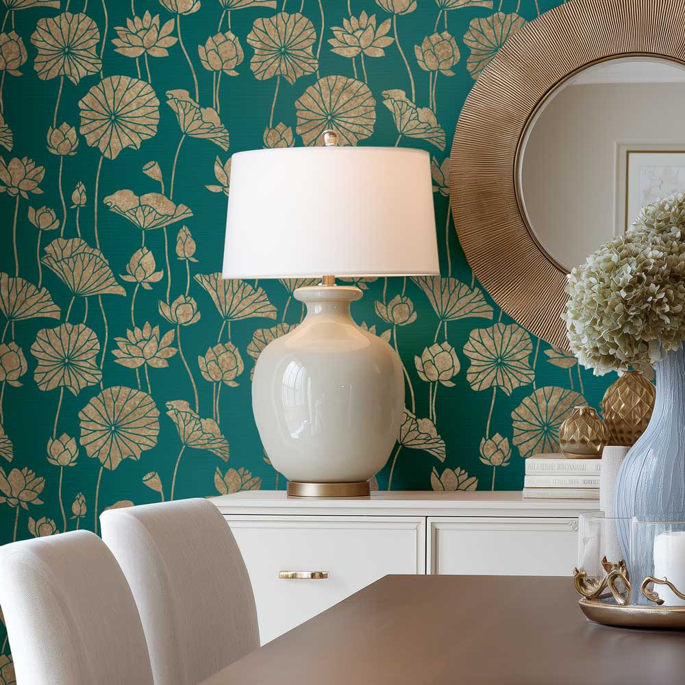 A white ceramic table lamp with a white drum shade and gold base sits on a white sideboard against a teal stenciled wall with a gold lotus floral pattern. A round wood-framed mirror hangs on the wall, and a dining table with white chairs is visible in the foreground.