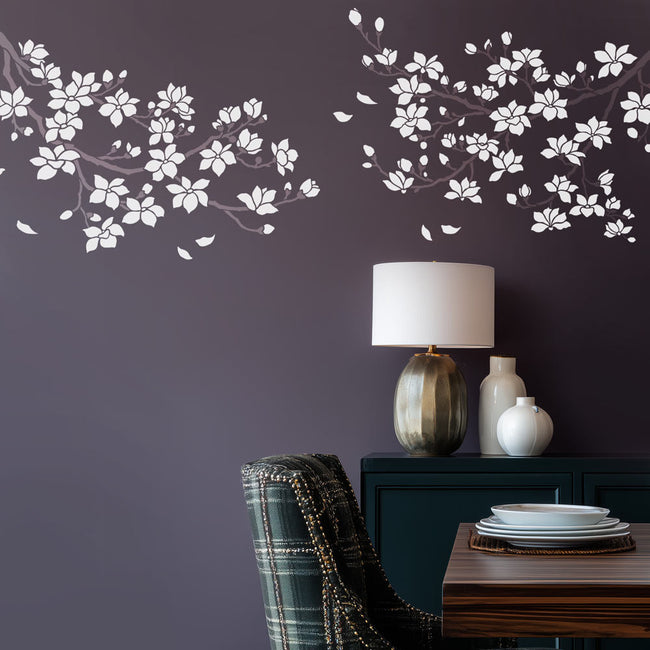 Decorative wall stencil of white floral branches on a dark purple wall with a lamp and vases on a table.